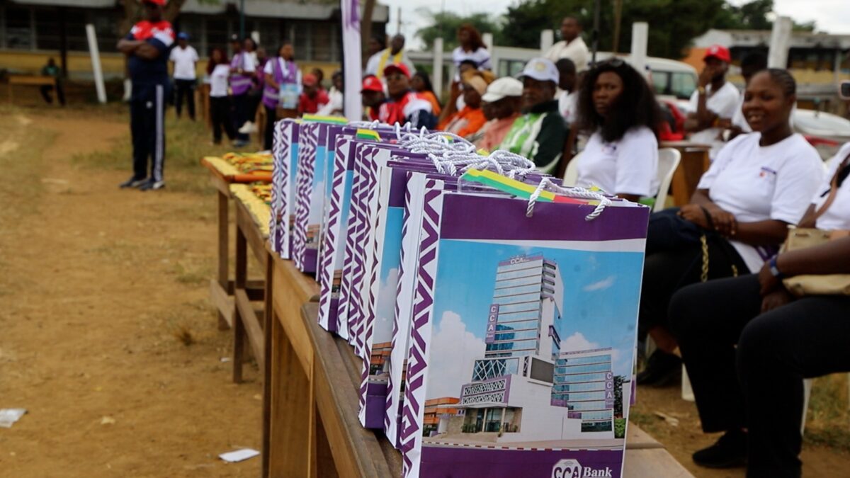 Une rangée de sacs de marque CCA-BANK est posée sur une table au premier plan. Les sacs sont blancs avec un motif géométrique violet et une image du siège de la banque, ainsi que le logo de l'institution. Ils ont des poignées blanches et des rubans aux couleurs nationales du Cameroun (vert, rouge, jaune). L'arrière-plan montre un public assis, légèrement flou.